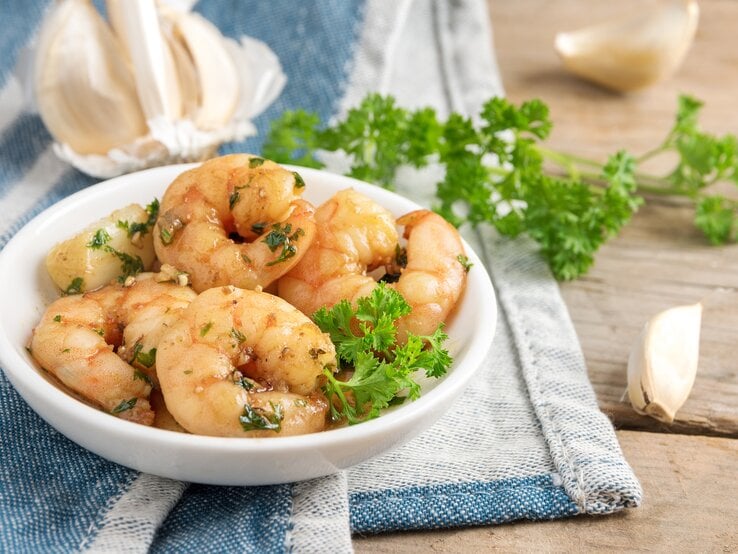 Mehrere Gambas von Ajo liegen in einer weißen Schüssel auf einem Geschirrhandtuch auf einem Holztisch. Um die Schüssel herum liegen Knoblauch und Petersilie.