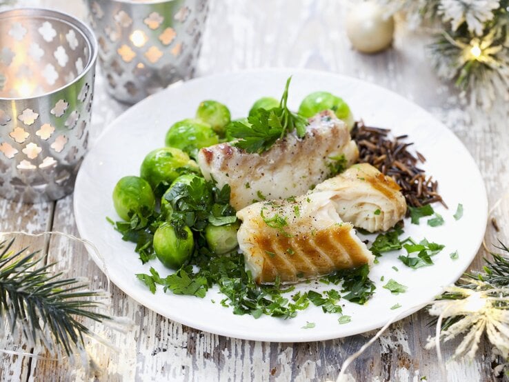 Ein festlich dekoriertes Tellergericht mit gebratenem Fisch, frischen Rosenkohl und Wildreis, garniert mit frischen Kräutern, auf einem rustikalen Holztisch. Im Hintergrund sind weihnachtliche Dekorationen wie Zweige und Kerzen zu sehen.