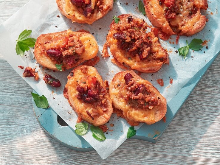 Das Bild zeigt mehrere kleine Scheiben Brot, die mit einer herzhaften Mischung aus geschmolzenem Belag, Bohnen und gehacktem, angebratenem Gemüse oder Fleisch belegt sind. Die Brotscheiben liegen nebeneinander auf einem mit Backpapier ausgelegten Brett. Zwischen den Broten liegen vereinzelt frische Kräuterblätter, die das Gesamtbild abrunden. Einige Krümel des Belags sind auf dem Backpapier verstreut.