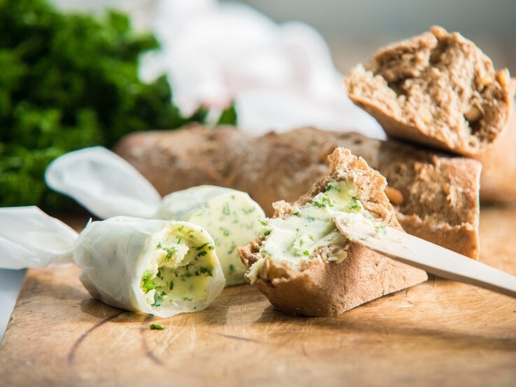 Hausgemachte Kräuterbutter eingewickelt in Brotpapier, angeschnitten auf einem Holzbrett. Daneben mehrere Scheiben Weißbrot.