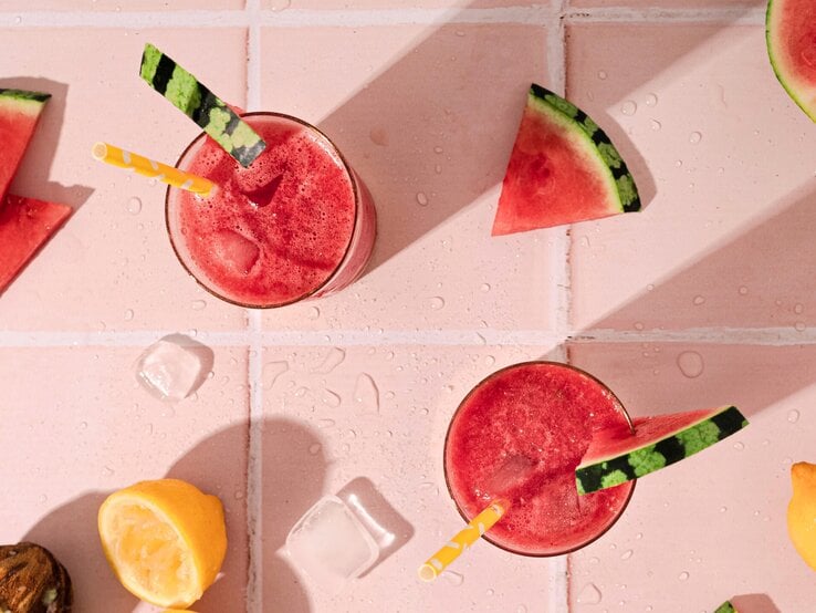 Wassermelone Frosé in zwei großen Gläsern mit Strohhalm und frischer Melone auf einem rosa Tisch aus Fliesen. 