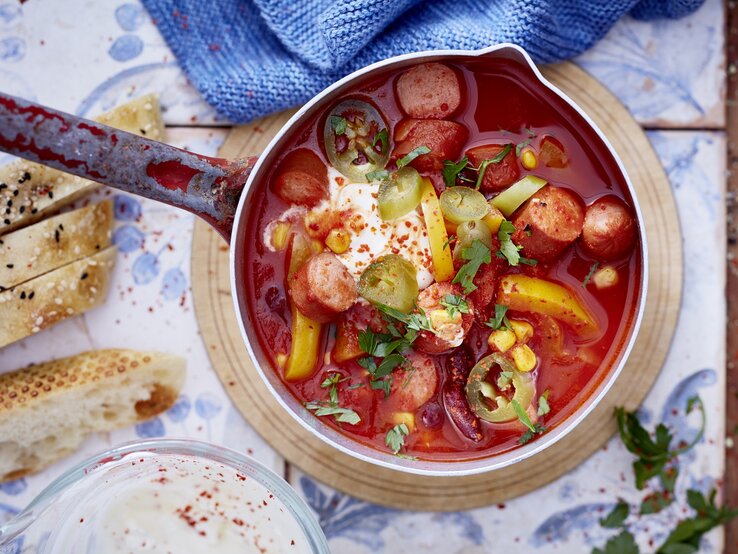 Ein Topf mit kräftig roter Suppe, gefüllt mit geschnittenen Würstchen, Mais, Kidneybohnen, Paprika, Jalapeños und einem Klecks Sauerrahm, garniert mit frischen Kräutern. Daneben liegen aufgeschnittenes Fladenbrot und ein Glas mit einer hellen Sauce. Der Topf steht auf einem runden Holzbrett auf einem gemusterten Untergrund.