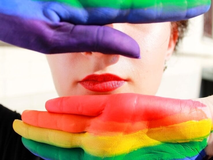 Junge Frau mit roten Lippen, deren Gesicht teils von zwei bunt bemalten Händen in Regenbogenfarben verdeckt wird.
