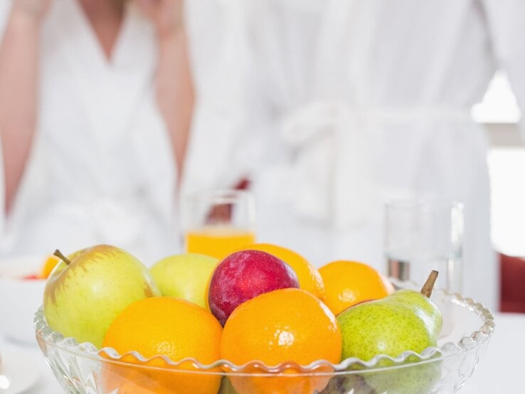 Obstschale mit Äpfeln, Birne, Pflaume und Mandarinen vor zwei Erwachsenen in weißen Bademänteln am Frühstückstisch.
