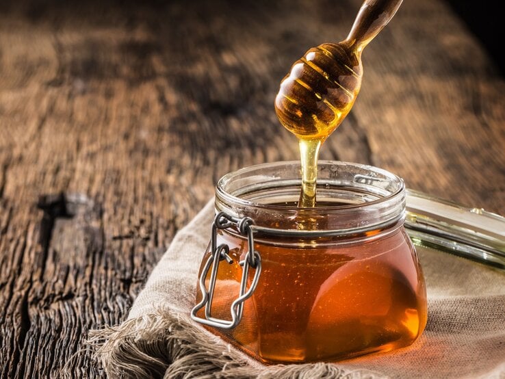 Ein Glas Honig mit Metallverschluss steht auf rustikalem Holz, ein Honiglöffel zieht goldenen Honig in die Höhe. | © Adobe Stock / weyo
