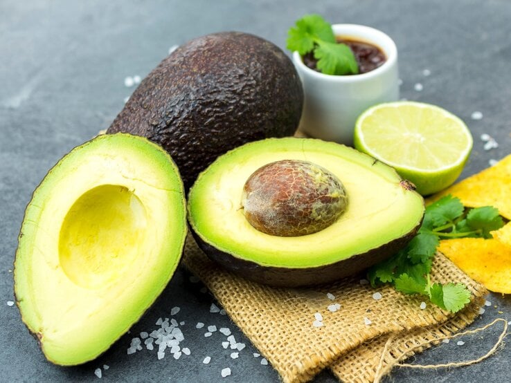 Aufgeschnittene Avocado mit Kern liegt neben Limettenhälfte, Koriander und Salz auf rustikalem Jutestoff. | © Adobe Stock / Jan