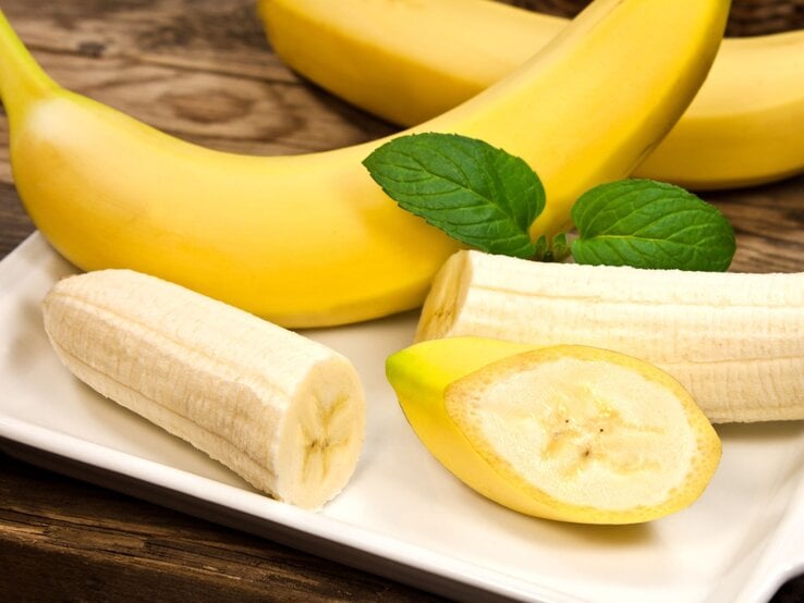 Frische Bananen, teils geschält und geschnitten, liegen dekorativ mit Minzblättern auf einem weißen Teller. | © Adobe Stock / PhotoSG