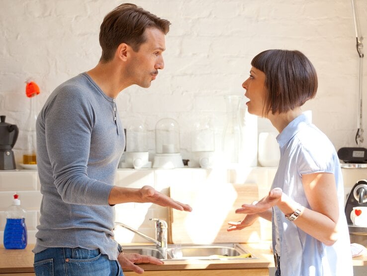 Paar, das in einer hellen Küche steht und eine hitzige Diskussion führt. Der Mann, gekleidet in ein graues Langarmshirt und Jeans, gestikuliert mit seinen Händen, während er offenbar etwas erklärt oder betont. | © iStock/anzeletti 