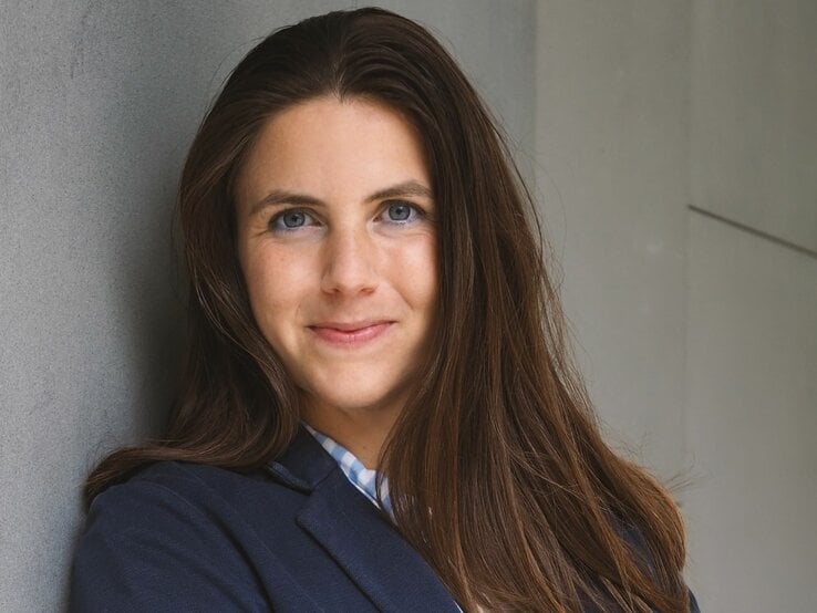 Frau mit langen, braunen Haaren und blauen Augen. Sie trägt ein dunkelblaues Jackett über einer karierten Bluse und lehnt an einer grauen Wand. Die Frau lächelt leicht und schaut direkt in die Kamera. | © privat