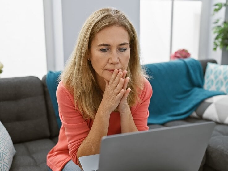 Eine nachdenkliche ältere Frau mit langen blonden Haaren sitzt im Wohnzimmer und blickt konzentriert auf ihren Laptop.