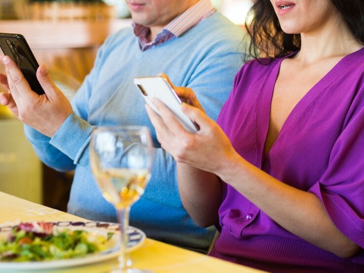 Eine Frau in lila Kleid und ein Mann in blauem Pulli starren beim Essen nebeneinander auf ihre Smartphones, ohne sich anzuschauen.