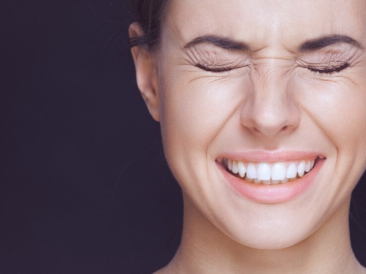 Frau, die die Augen fest geschlossen hat und lächelt. Man sieht ihre weißen Zähne, und sie scheint entweder zu lachen oder einen intensiven Moment zu erleben.