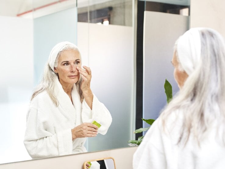 Frau im Bademantel steht vor dem Spiegel und trägt Augencreme auf 