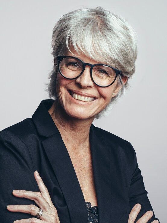 Ältere Frau mit silbergrauem Pixie-Cut, schwarzem Blazer und Brille lächelt selbstbewusst mit verschränkten Armen vor grauem Hintergrund. | © Shutterstock/Jacob Lund