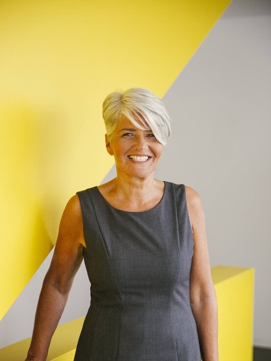 Lachende ältere Frau mit silbergrauem Kurzhaarschnitt in grauem Etuikleid steht vor gelber Wand mit grafischem Muster. | © imago/Westend61