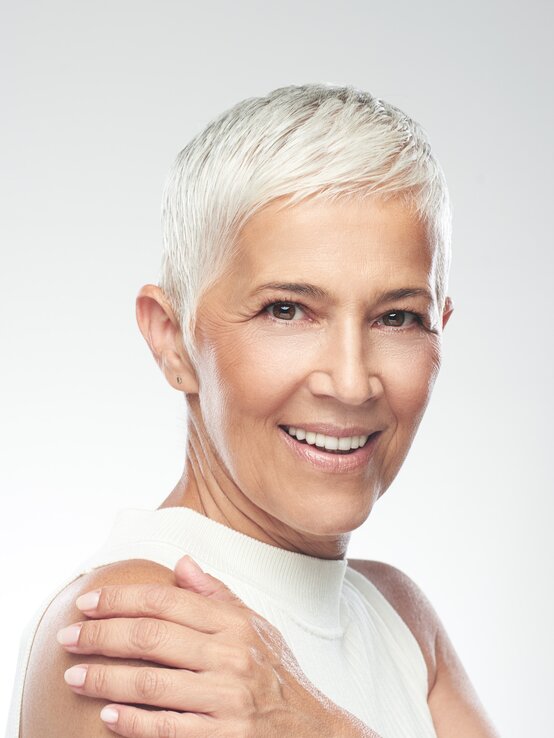 Ältere Frau mit kurz geschnittenem weißgrauem Haar, ärmellosem Top und gepflegter Haut lächelt mit der Hand auf ihrer Schulter. | © millann/Getty Images