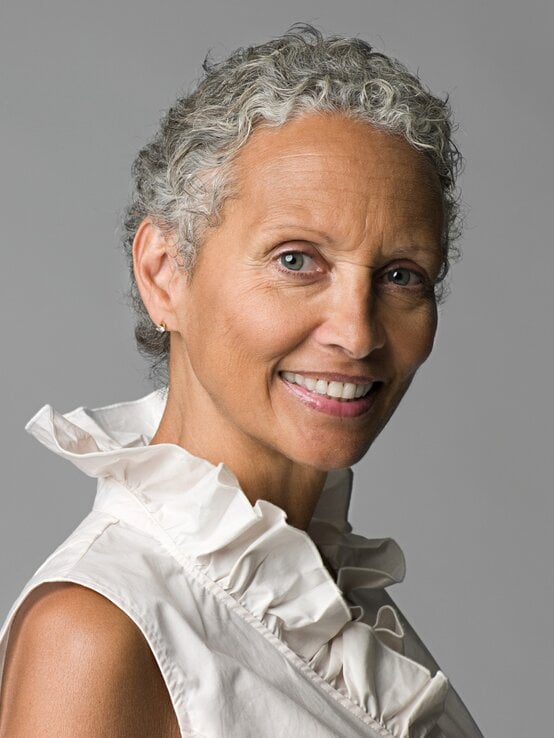 Ältere Frau mit kurzem lockigem Grauhaar und heller Rüschenbluse lächelt freundlich | © Shutterstock/Image Source Collection