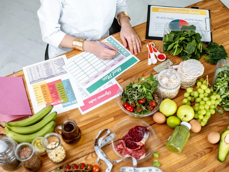 Ernährungsberaterin erstellt einen Ernährungsplan am Tisch, umgeben von frischen Lebensmitteln wie Obst, Gemüse, Fleisch, Nüssen und Vollkornprodukten sowie Unterlagen zu Makronährstoffen und einem Maßband.