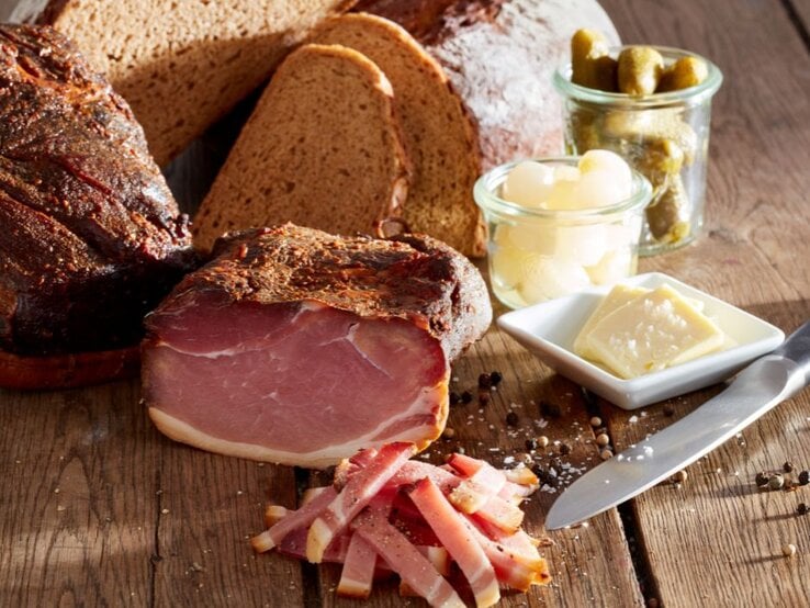 Rustikales Stillleben mit aufgeschnittenem geräuchertem Schinken auf einem Holztisch, dazu in Scheiben geschnittenes Bauernbrot, ein Messer, Gewürze sowie kleine Gläser mit Essiggurken und Perlzwiebeln und ein Tellerchen mit Butterstückchen.