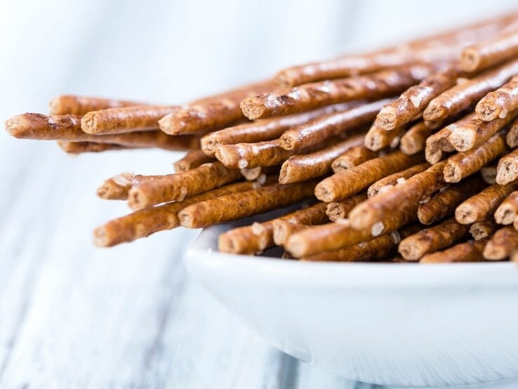 Ein weißer Teller mit einer Ansammlung dünner Salzstangen, die im Vordergrund liegen. Der Fokus liegt auf den Salzstangen und ihre leicht körnige Salzstruktur ist sichtbar. Der Hintergrund ist hell und unscharf.