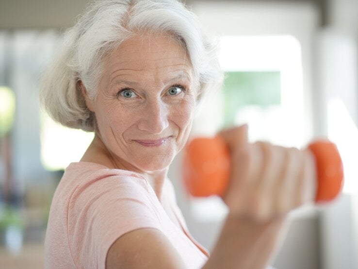 Ältere Frau mit grauem Haar und rosa T-Shirt hält eine orangefarbene Hantel und lächelt motiviert in die Kamera. Sie scheint sich in einem hellen, modernen Raum zu befinden und wirkt aktiv und gesund.