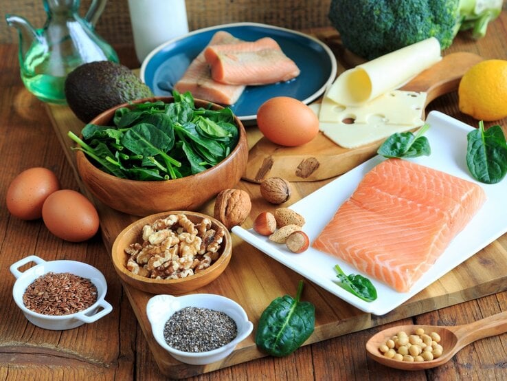 Eine Auswahl magnesiumreicher Lebensmittel auf einem rustikalen Holztisch. Im Vordergrund frischer Lachs auf einem weißen Teller, daneben eine Schale mit Walnüssen, Chia- und Leinsamen. Zusätzlich enthalten: frischer Spinat in einer Holzschale, Eier, Avocado, verschiedene Nüsse, Käse, Brokkoli, Zitronen und eine Karaffe mit Öl. | © Adobe Stock/  Juefrateam