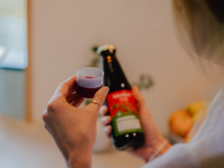 Junge Frau mit blondem Haar hält Messbecher mit dunkelrotem Saft EisenPlus in der einen und eine Flasche mit rotem Etikett in der anderen Hand. | © Rabenhorst