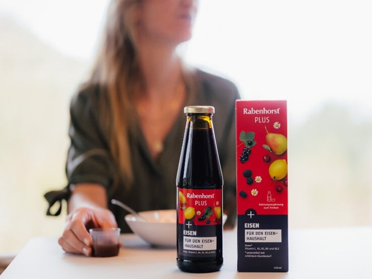 Nahaufnahme einer Nahrungsergänzung mit Eisen: Rabenhorst PLUS vor unscharfer Frau, die den Saft am Tisch trinkt. | © Rabenhorst