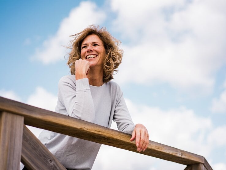 Fröhliche Frau mittleren Alters blickt verträumt in die Ferne, während sie sich an ein Geländer unter wolkigem Himmel lehnt.