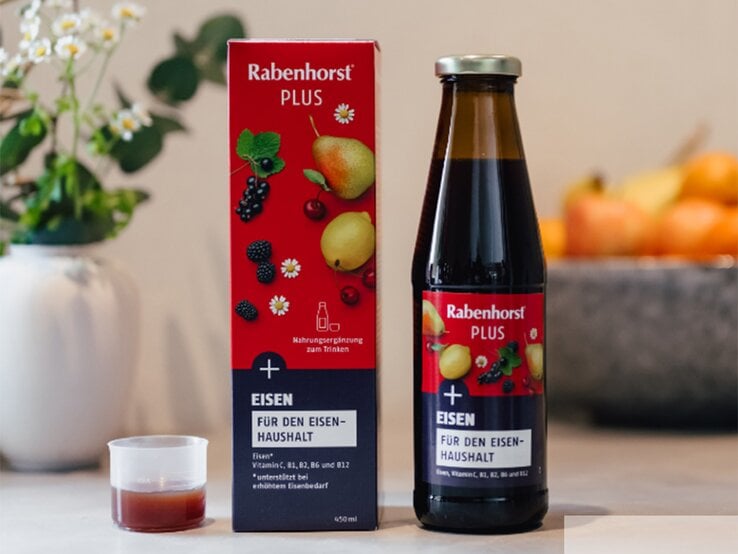 Rabenhorst PLUS Eisen in Glasflasche und roter Verpackung auf hellem Tisch mit Dekovase und Schale voller Äpfel im Hintergrund. | © Rabenhorst
