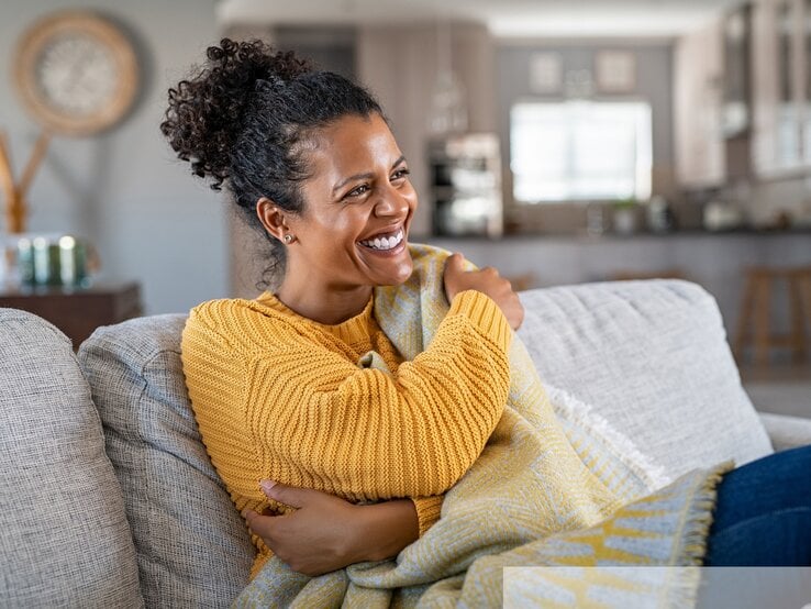 Fröhliche junge PoC Frau mit braunen, lockigen Haaren und gelbem Pullover, die sich entspannt in eine Decke gehüllt auf ihrem Sofa zurücklehnt. | © stock.adobe.com/Rido