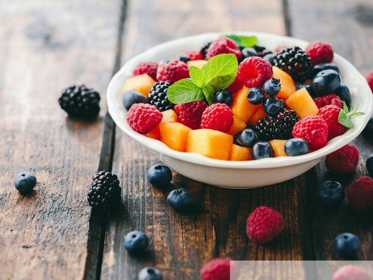 Eine weiße Schale mit frischen Beeren wie Himbeeren, Heidelbeeren und schwarzen Brombeeren, dazu Orangenstücke und ein grünes Minzblatt auf rustikalem Holztisch. | © stock.adobe.com/Melica