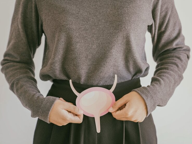 Eine Frau in einem grauen Pullover hält ein rosafarbenes, abstrahiertes Modell einer Blase vor ihrem Bauch. | © stock.adobe.com/SewcreamStudio