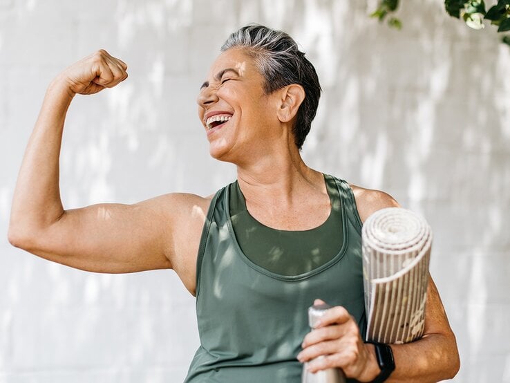 Eine lächelnde ältere Frau mit kurzen grauen Haaren zeigt stolz ihre Muskeln und hält eine Wasserflasche und Yogamatte in der Hand. | © stock.adobe.com/ (JLco) Julia Amaral