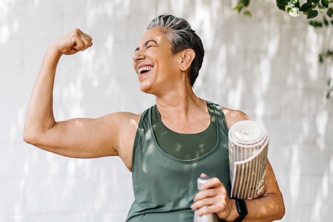 Eine lächelnde ältere Frau mit kurzen grauen Haaren zeigt stolz ihre Muskeln und hält eine Wasserflasche und Yogamatte in der Hand. | © stock.adobe.com/ (JLco) Julia Amaral