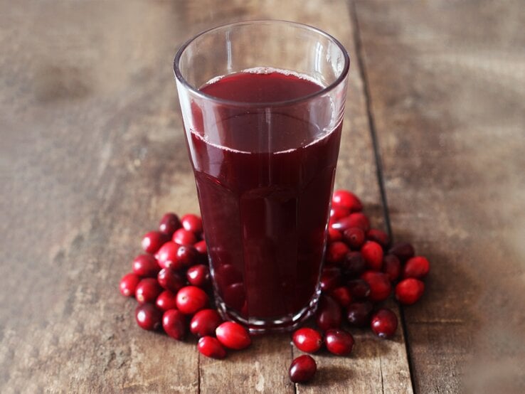 Ein Glas dunkler Cranberrysaft steht auf rustikalem Holztisch, umgeben von frischen roten Cranberries. | © stock.adobe.com/Printemps