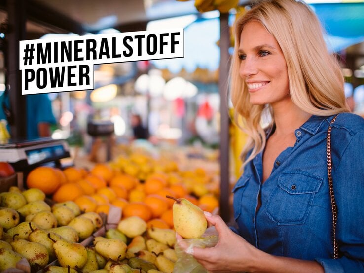 Junge blonde Frau in Jeanshemd lächelt zufrieden beim Auswählen von Birnen an einem bunten Obststand auf dem Wochenmarkt.
