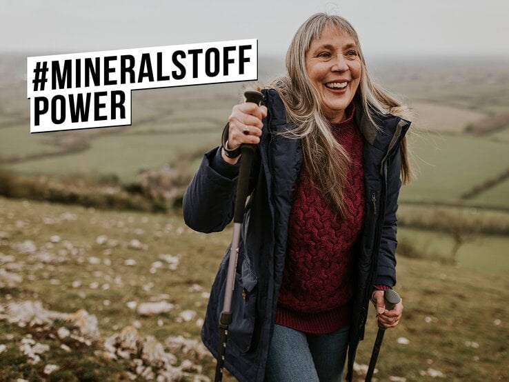 Lachende ältere Frau mit langen grauen Haaren wandert mit Stöcken im Wollpullover und Winterjacke über grünen Hügel.