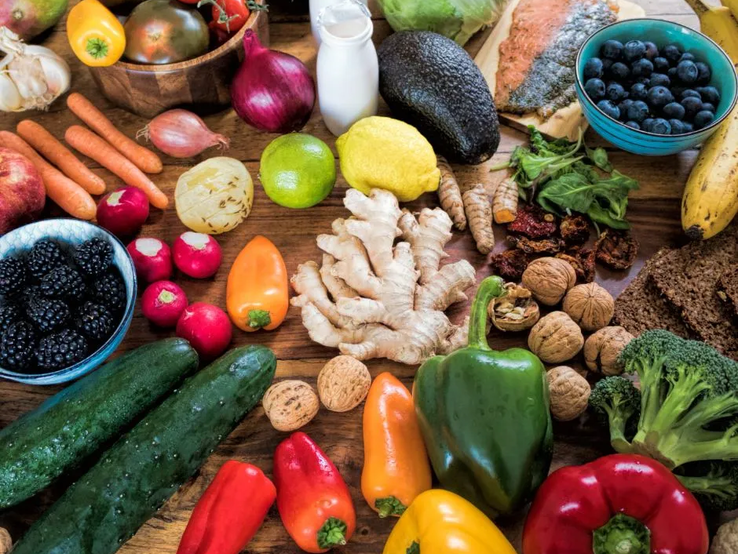 Ein Holz-Tisch mit einer Vielzahl von gesunden Lebensmitteln, darunter frisches Obst wie Zitronen, Limetten, Bananen, Blaubeeren und Brombeeren, Gemüse wie Paprika, Brokkoli, Gurken, Karotten, Rettich und Avocado, sowie weitere Zutaten wie Ingwer, Walnüsse, getrocknete Tomaten, Knoblauch, Vollkornbrot und Lachs. Im Hintergrund sind auch kleine Glasflaschen mit einer Milch- oder Joghurt-ähnlichen Flüssigkeit zu sehen.