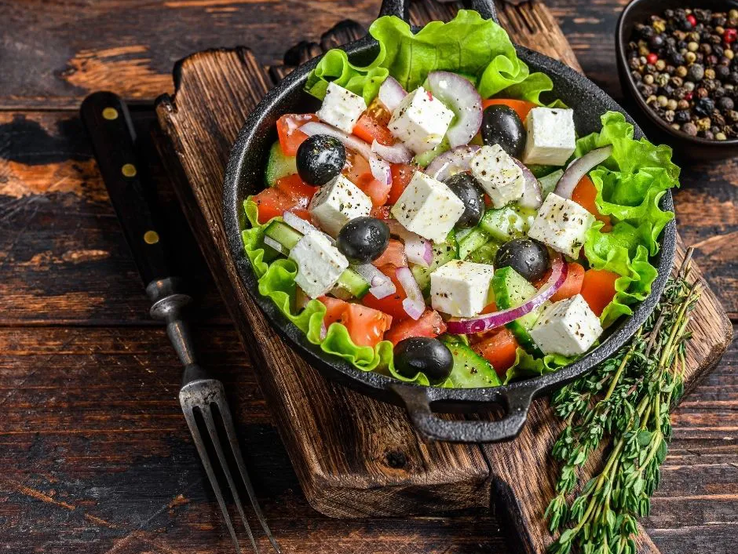 Ein frischer griechischer Salat in einer schwarzen Pfanne, serviert auf einem rustikalen Holzbrett. Der Salat besteht aus grünen Salatblättern, Tomaten, Gurkenscheiben, roten Zwiebelringen, schwarzen Oliven und gewürfeltem Feta, der leicht mit Kräutern bestreut ist. Im Hintergrund steht eine Schale mit buntem Pfeffer. Daneben liegt eine Gabel sowie ein Bund frischer Kräuter, vermutlich Thymian. Der Untergrund ist eine rustikale Holzoberfläche.