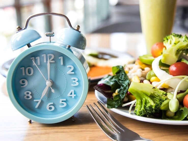 Eine hellblaue Weckeruhr auf einem Holztisch neben einem Teller mit frischem Salat. Der Salat ist bunt und beinhaltet Gemüse wie Brokkoli, Tomaten, Mais und grüne Bohnen. Im Hintergrund ist verschwommen eine weitere Mahlzeit und ein Getränk zu sehen.