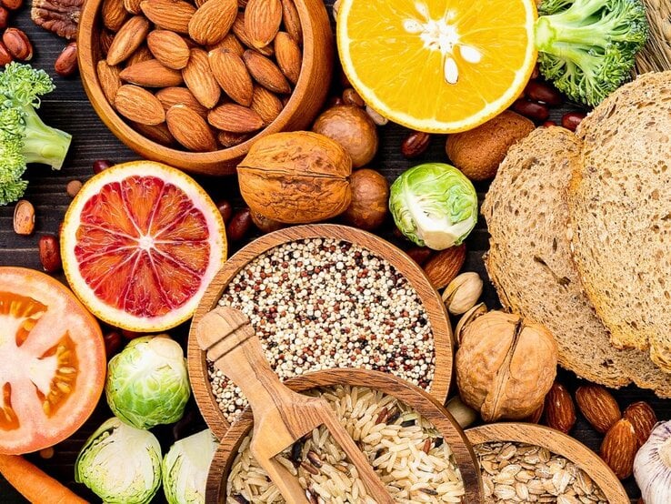 Verschiedene ballaststoffreiche Lebensmittel wie Vollkornbrot, Nüsse, Samen, Reis, Quinoa, Hülsenfrüchte, Brokkoli, Rosenkohl, Karotten und Zitrusfrüchte liegen dekorativ auf einem dunklen Untergrund.