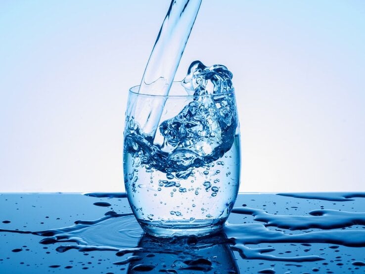 Klares Wasser wird in ein transparentes Glas gegossen, dabei spritzt es dynamisch über den Rand; das Glas steht auf einer nassen Oberfläche vor einem hellblauen Hintergrund. | © Getty Images/ K-Paul via Canva.com
