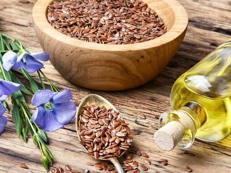 Holzschale mit Leinsamen, daneben eine Flasche Leinöl, ein Löffel mit Leinsamen und blühende blaue Leinpflanzen auf einem Holztisch.