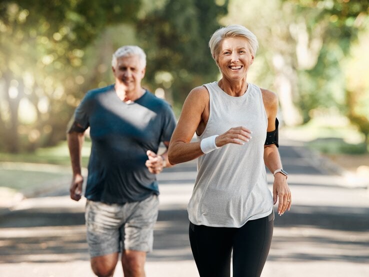 Älteres Paar, das gemeinsam im Freien joggt. Beide sehen glücklich und entspannt aus, während sie durch eine von Bäumen gesäumte Straße laufen.