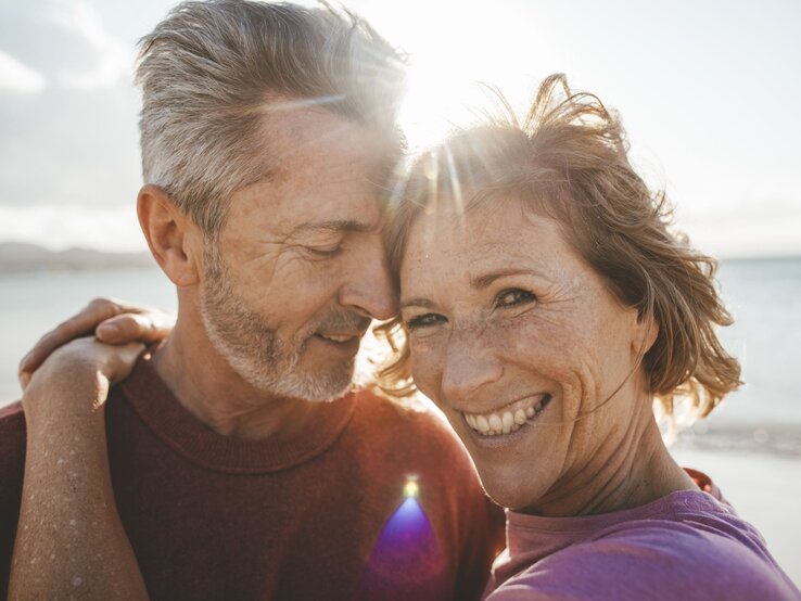 Eine Frau und ein Mann mittleren Alters umarmen sich im Sonnenschein. Die Frau lächelt unbeschwert in die Kamera.