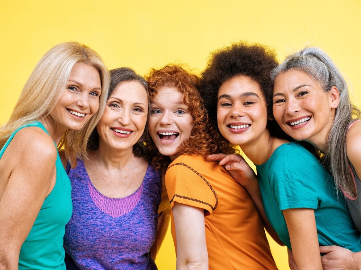 Fünf lachende Frauen verschiedener Altersgruppen stehen in bunter Kleidung eng zusammen vor gelbem Hintergrund. | © Adobe Stock / oneinchpunch