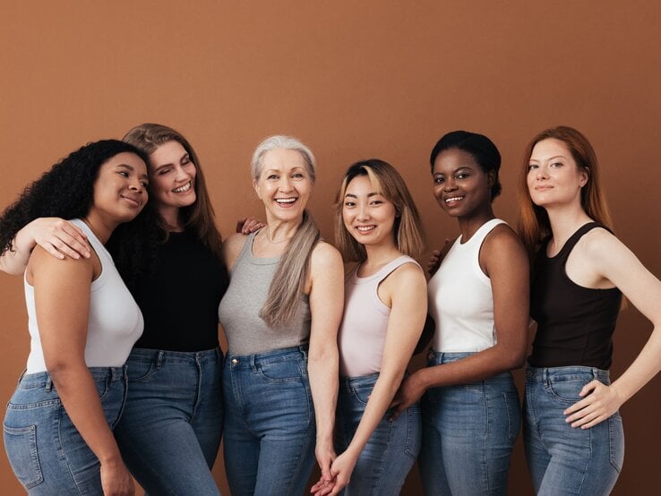 Sechs Frauen verschiedener Altersgruppen und Ethnien stehen eng umschlungen, lächelnd in Jeans und Tops vor braunem Hintergrund.