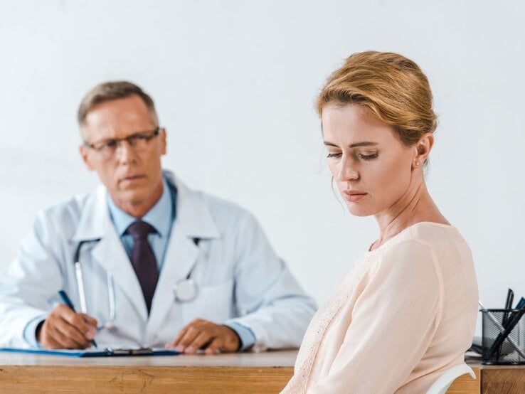 Eine nachdenklich blickende Frau sitzt mit gesenktem Kopf vor einem älteren Arzt in weißem Kittel an einem Schreibtisch.