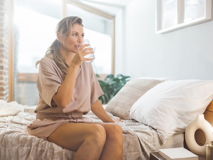 Eine Frau sitzt am Bettrand und trinkt aus einem Glas Wasser.